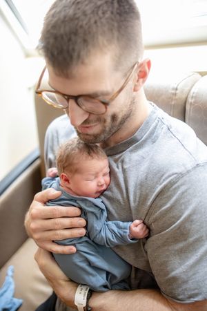 Father with glasses carefully cradling alert newborn baby dressed in blue in hospital room.