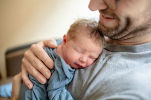 Close-up of bearded father holding sleeping newborn baby against his gray shirt.
