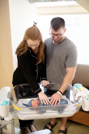New parents in hospital room lovingly looking at newborn baby lying in clear bassinet.