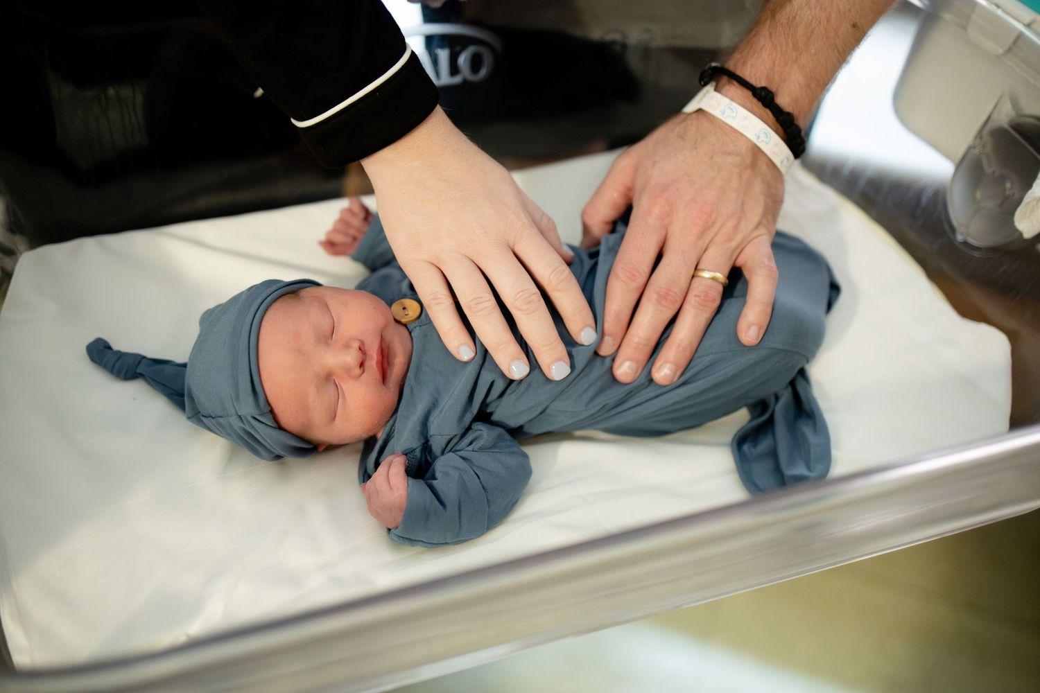 Overhead view of sleeping newborn baby in blue swaddle with parents hands resting gently on chest.