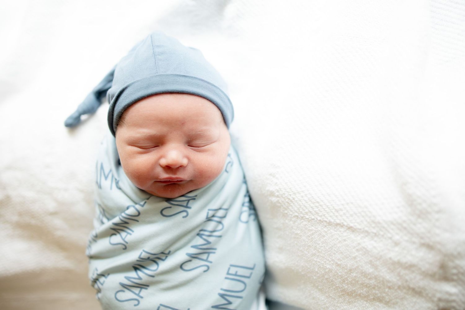 Sleeping newborn baby named Samuel swaddled in a blue personalized name blanket wearing a knotted hat.