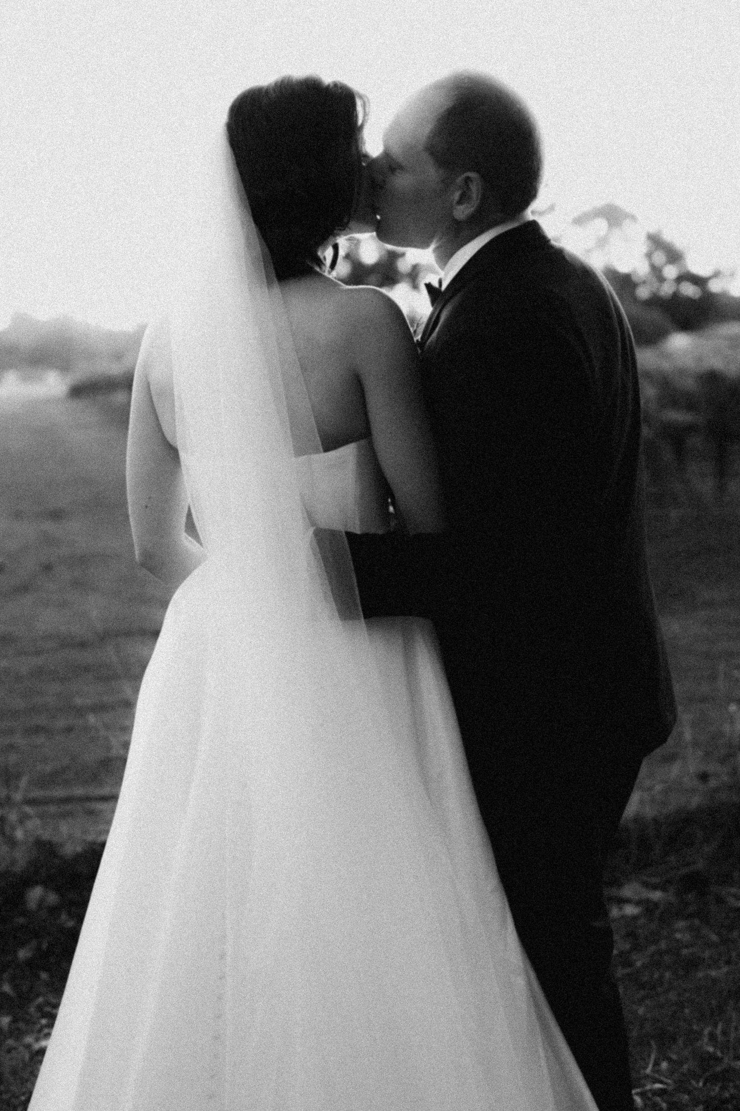 Black and white photo of a bride and groom sharing a kiss outdoors, bride wearing a flowing gown and veil.