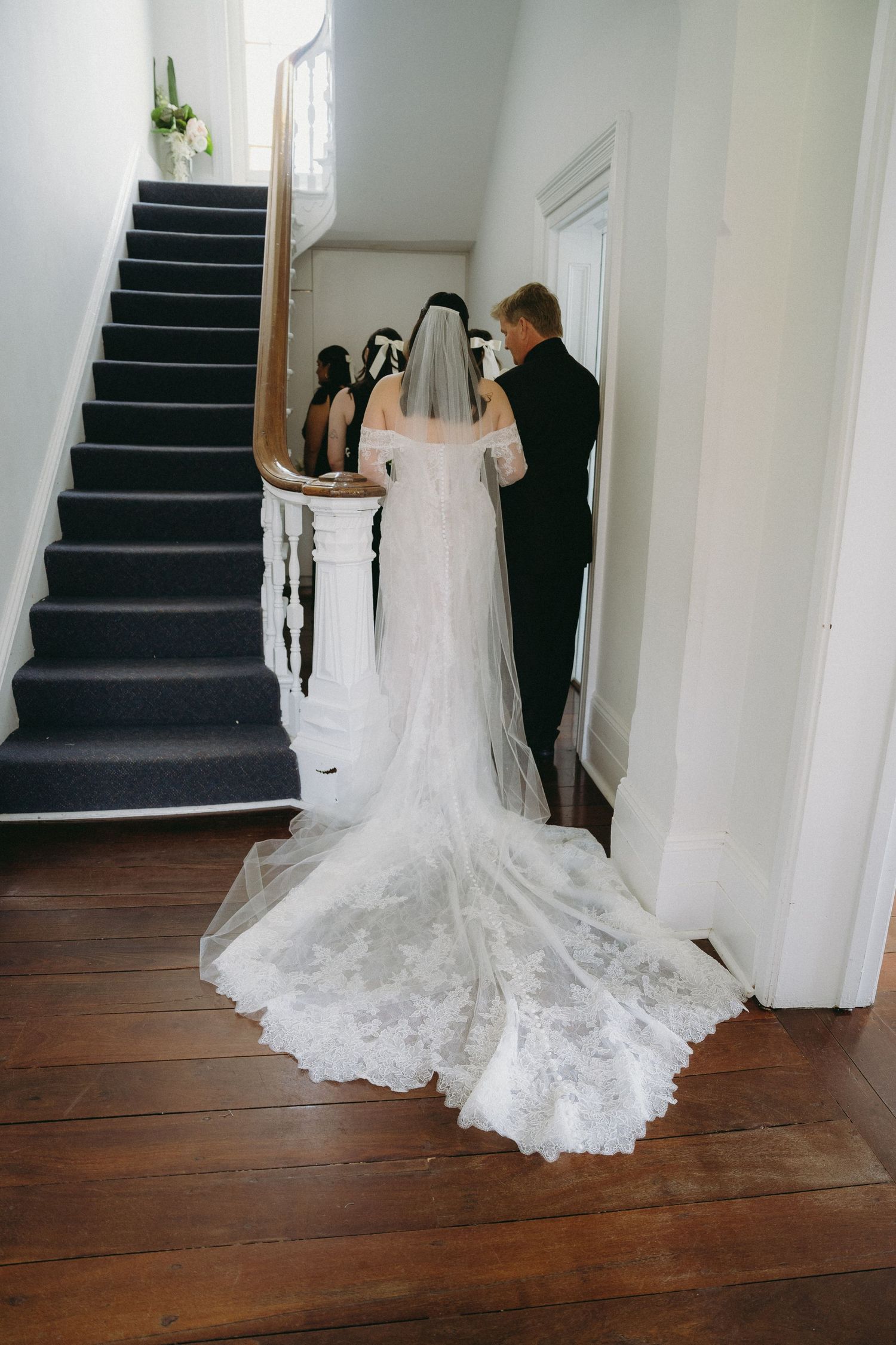 Bride in lace wedding gown with cathedral veil and train stands at staircase with groom in black suit.