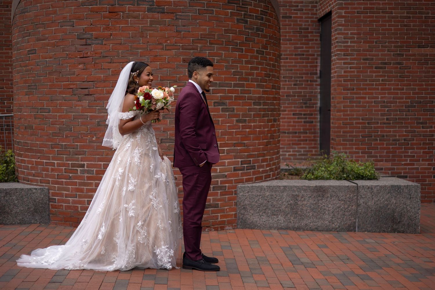 A bride in a lace wedding gown and groom in burgundy suit stand against a brick wall on their wedding day.