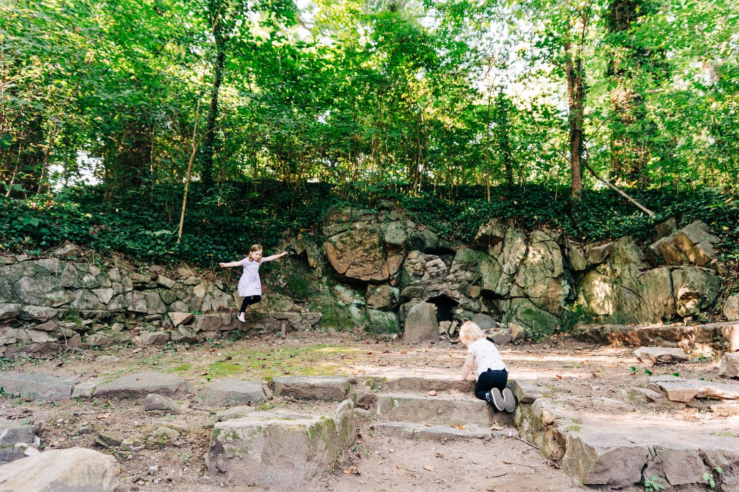 A series of stone ruins and walls nestled among lush green trees and vegetation in a forest setting where children are playing.