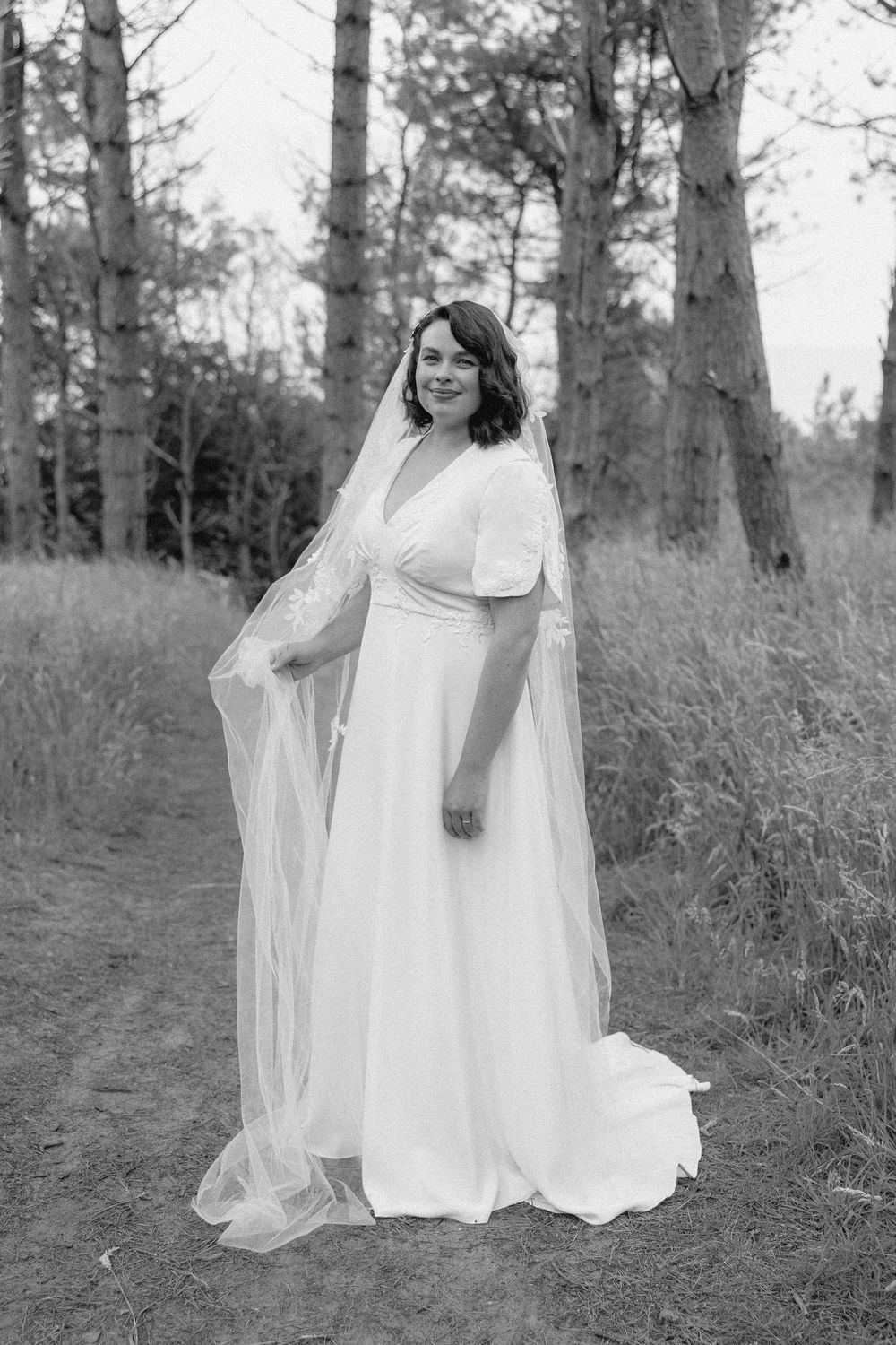 Black and white photo of a bride in an elegant white dress with flowing veil standing on a forest path.