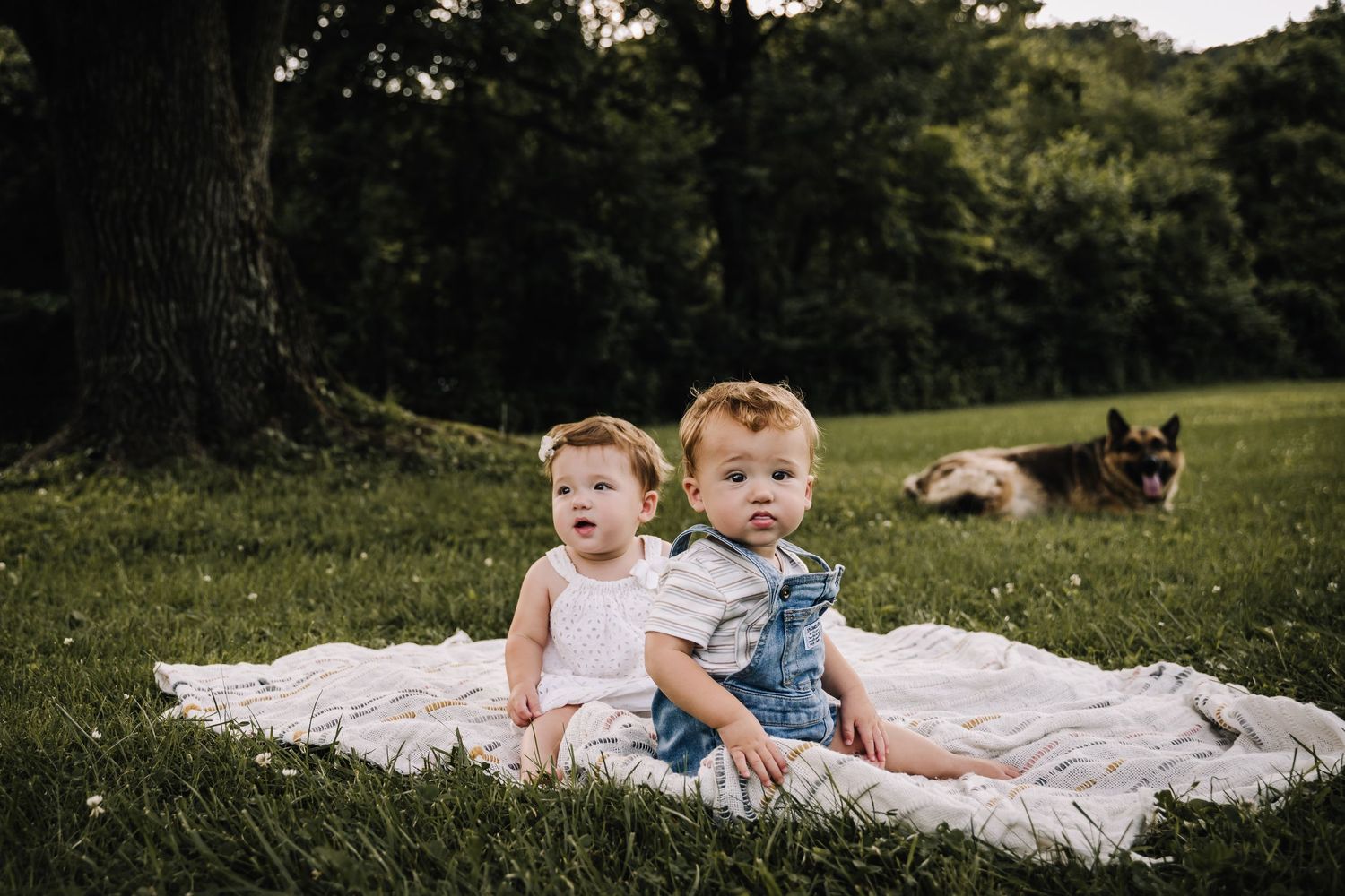 Two twin children in casual clothing lay on a white blanket in a grassy field during a sunny summer day. Family photographer in Middle TN