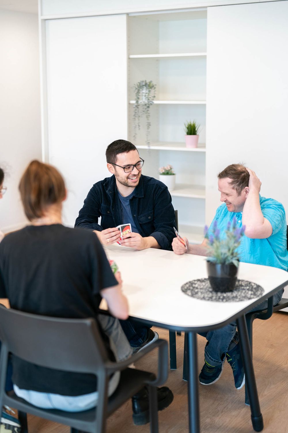 Several people sit at white tables in a modern office space with white shelving units and minimalist decor.