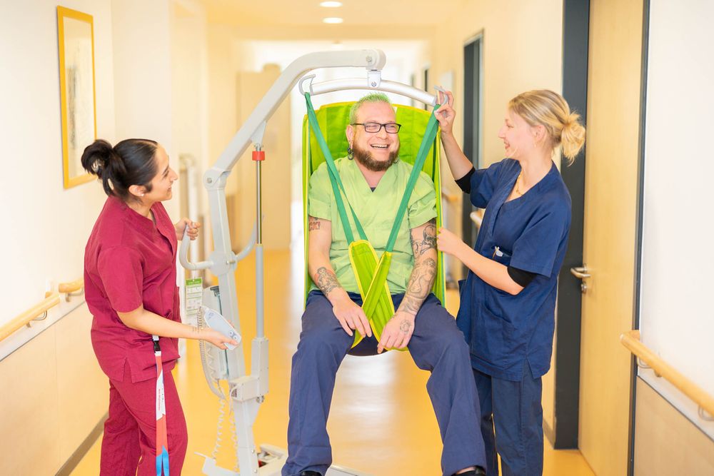 Healthcare workers in blue scrubs and high visibility vests assist patients in a bright hospital hallway.