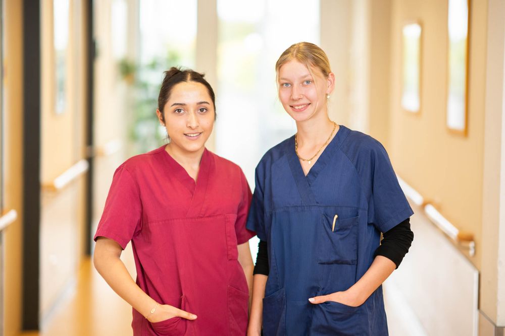Two healthcare workers in scrubs stand smiling together in a hospital hallway.