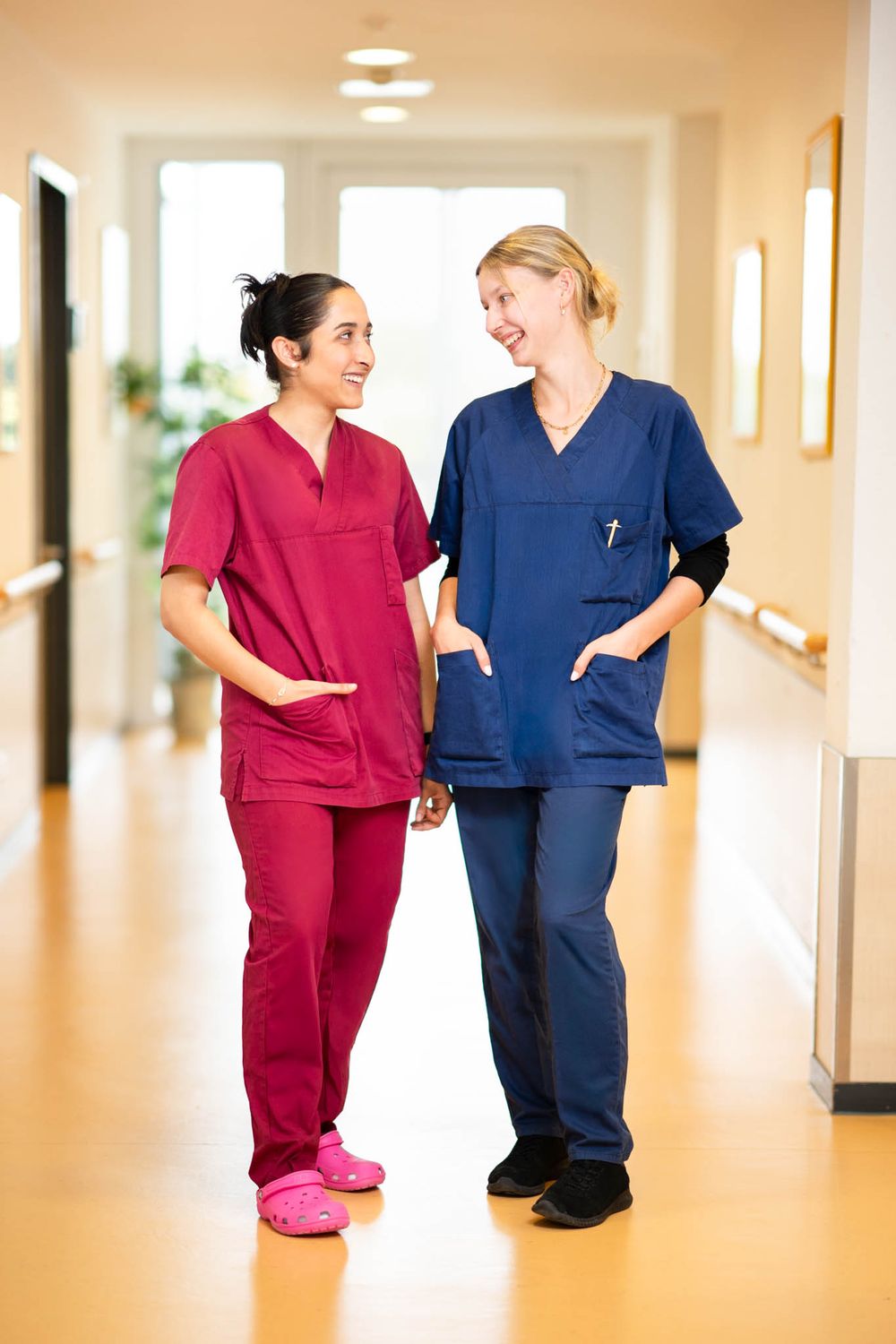 Healthcare workers in red and blue scrubs having a friendly conversation while walking down a bright hospital hallway.