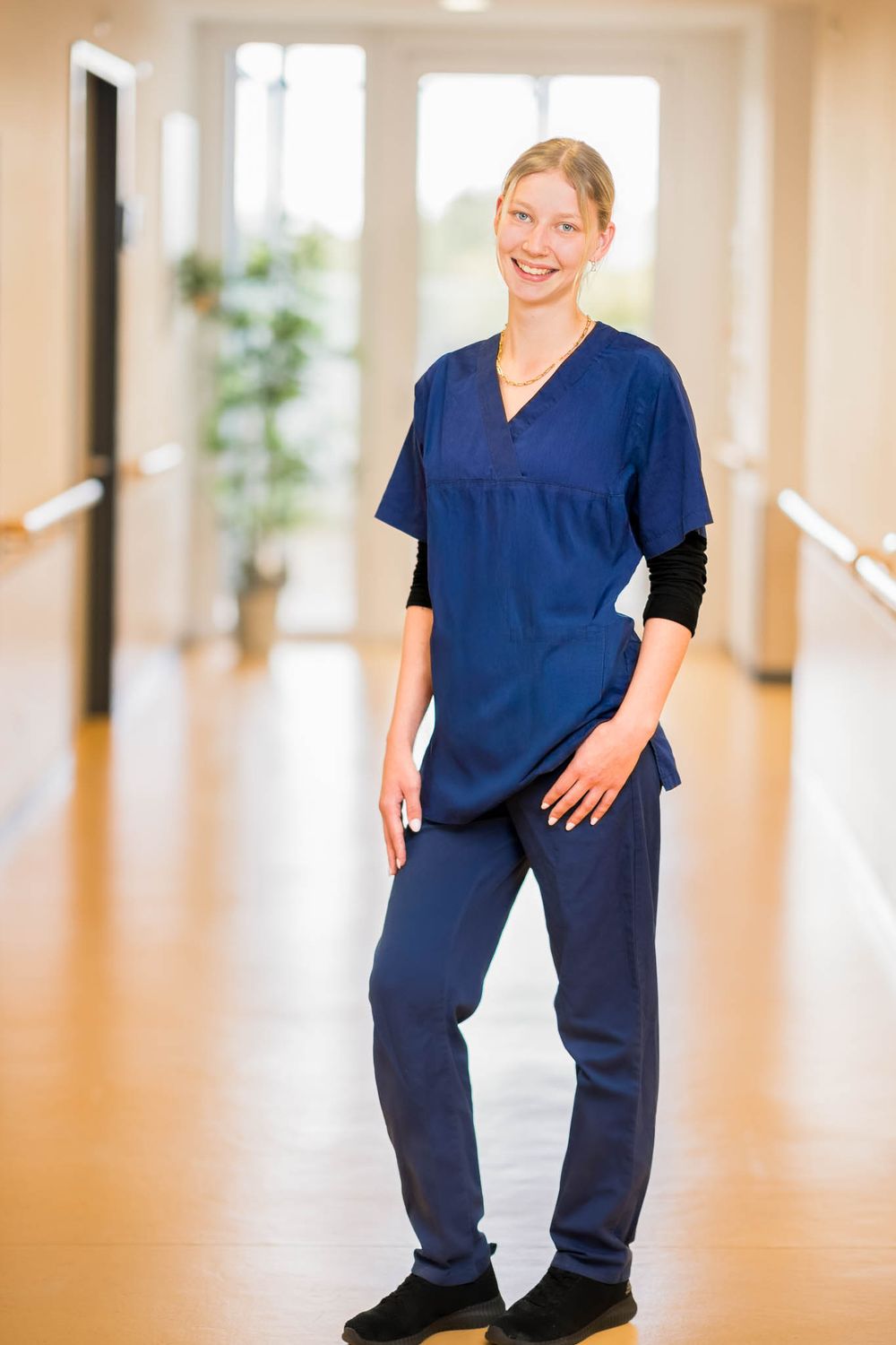 Healthcare worker in navy blue scrubs stands in a bright hospital hallway with a friendly expression.