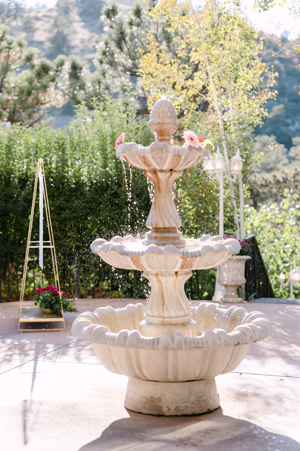 Elegant white stone fountain with tiered basins set against a lush garden backdrop with mountains in the distance.
