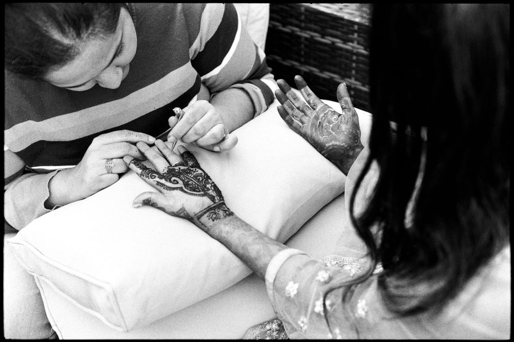 Black and white close-up of intricate henna mehndi being applied to a womans hand resting on a pillow.