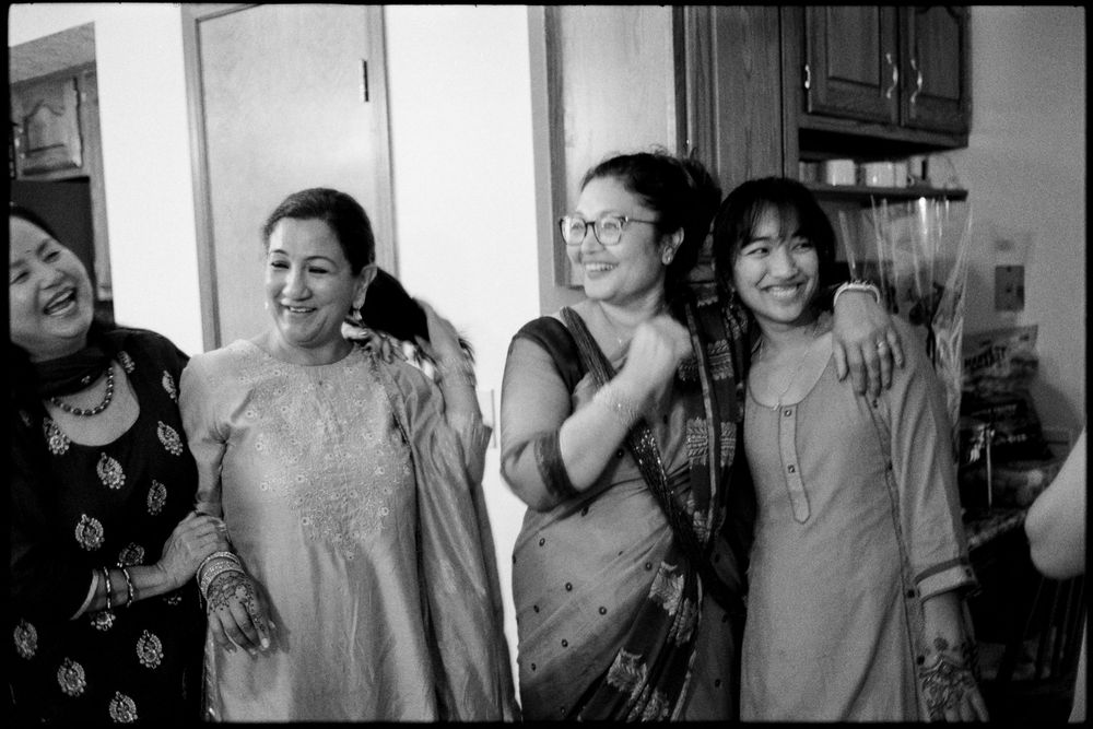Four women in traditional South Asian attire smile and embrace together in a kitchen in black and white.