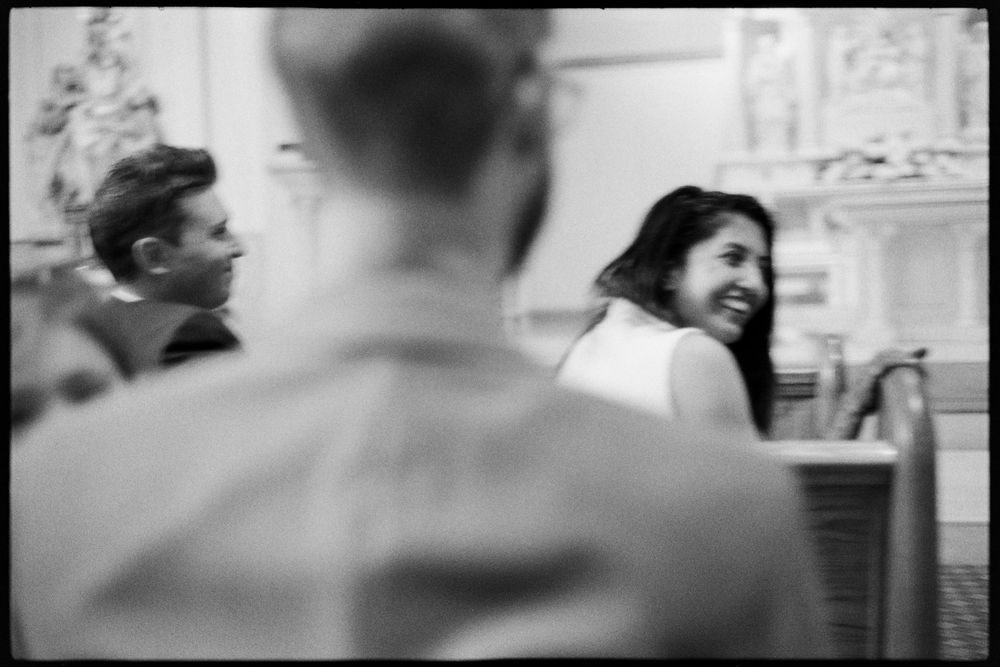 Black and white candid photo of a smiling woman seated in a church pew during a wedding ceremony.