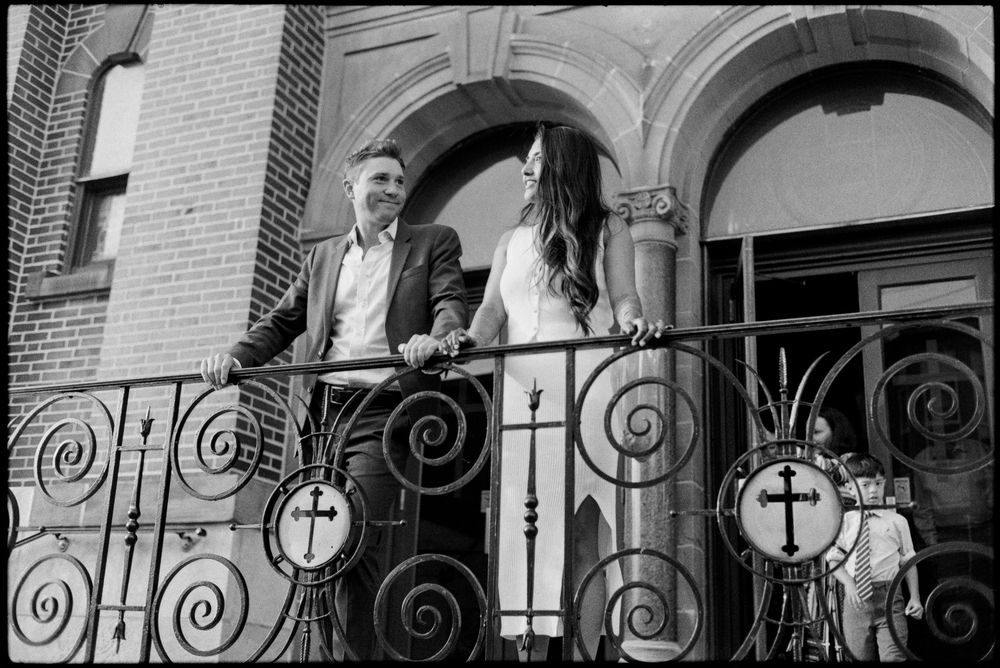 Black and white photo of a couple standing on an ornate iron balcony outside a brick church building.
