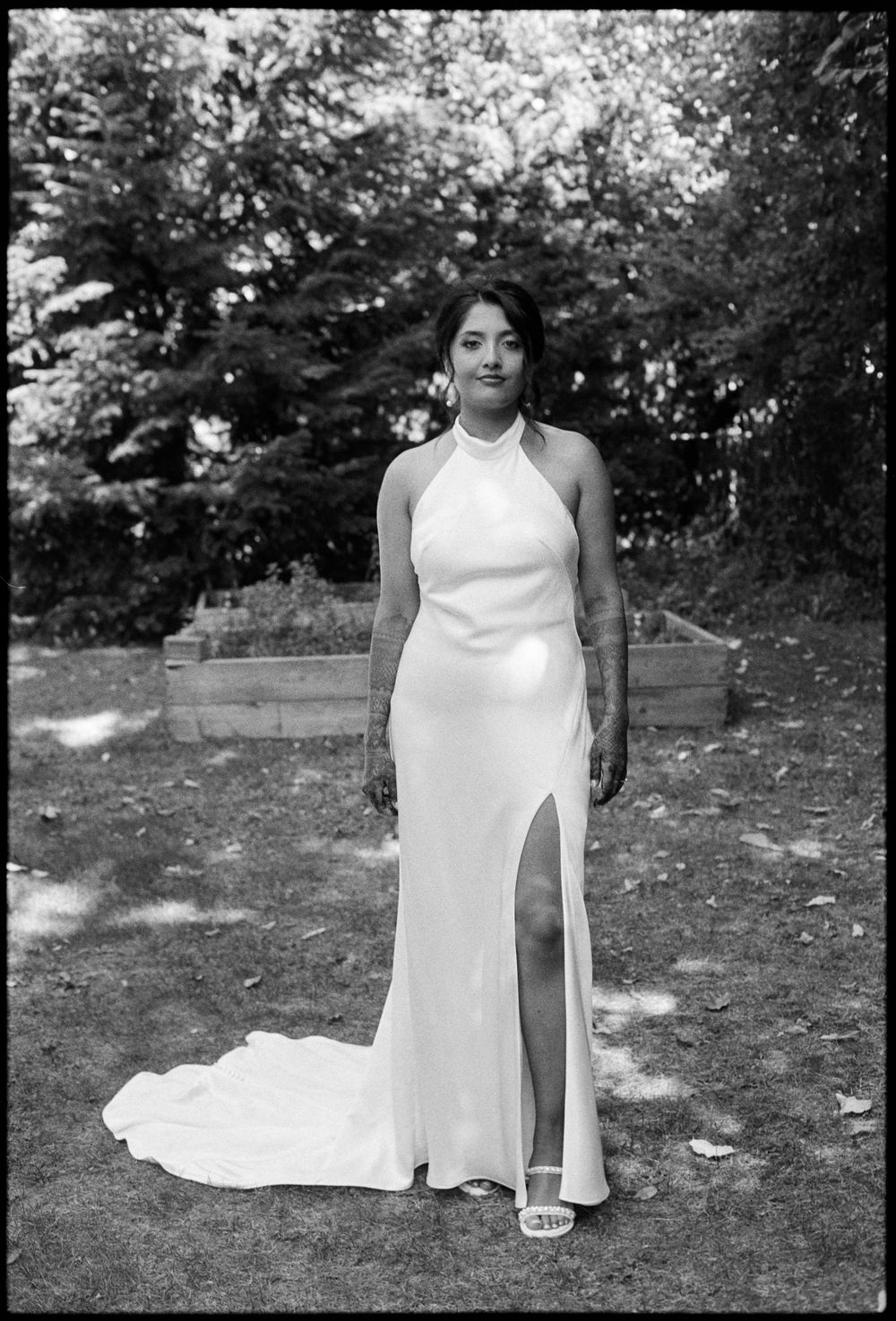 Black and white photo of a woman in a white halter neck gown with a slit, posing outdoors in a garden.
