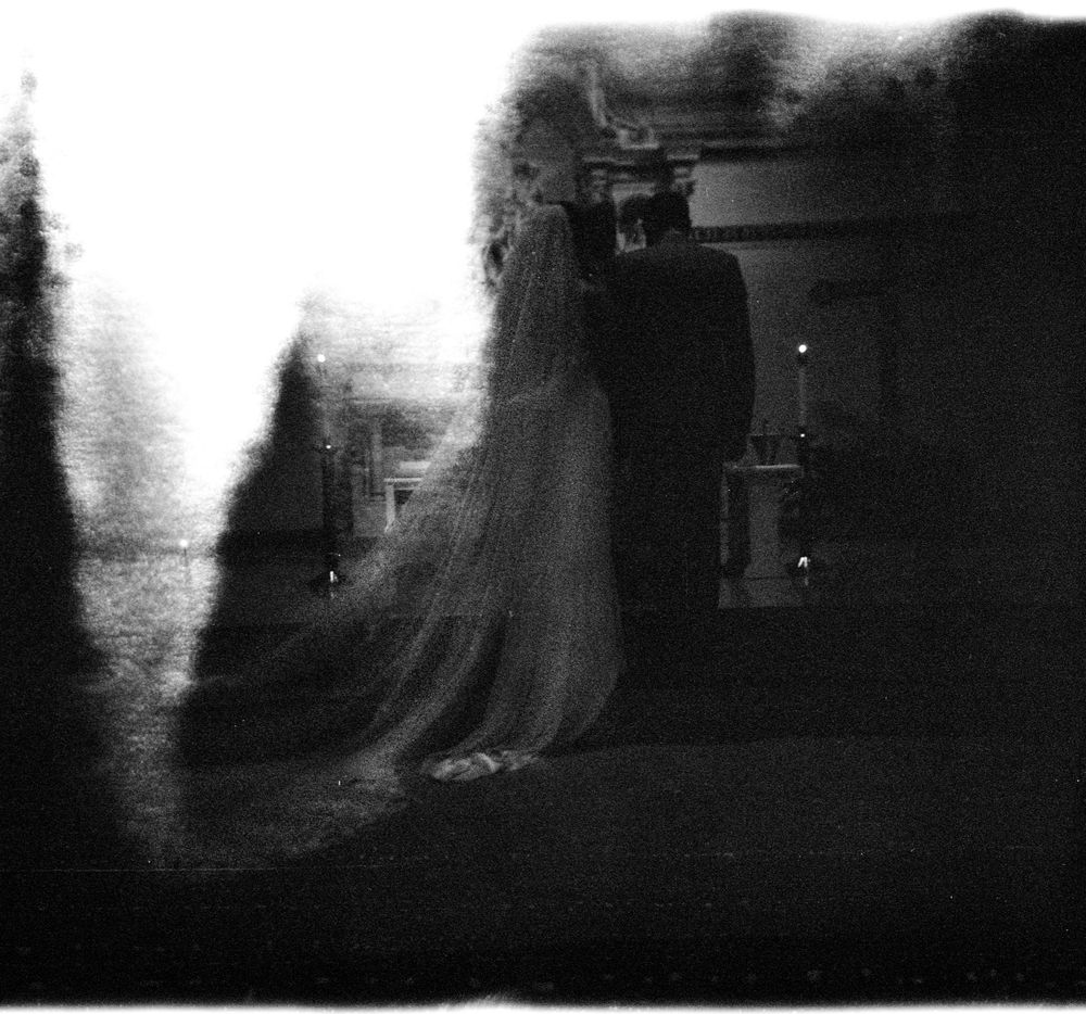 Ethereal black and white blurred image of bride in veil standing in a darkened church setting.