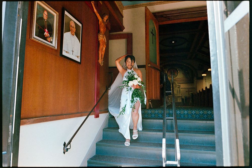 Bride in white gown carries floral bouquet up church steps past religious portraits and crucifix.