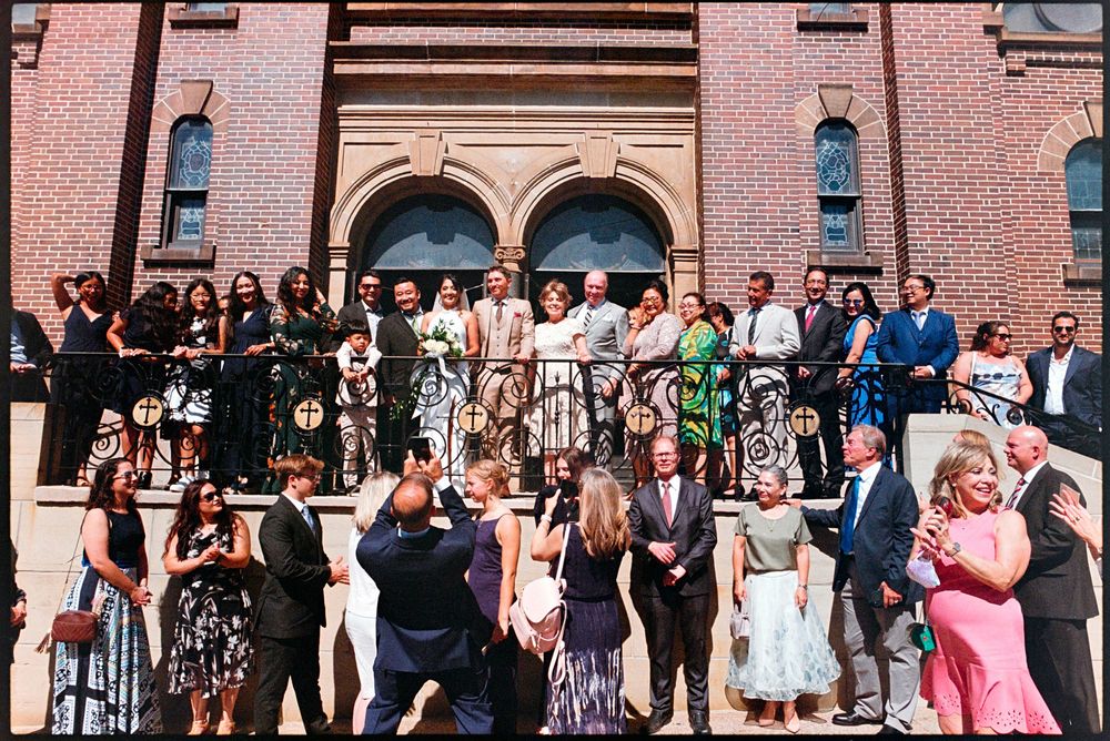 Large wedding party group photo on ornate brick church steps in sunny outdoor setting.