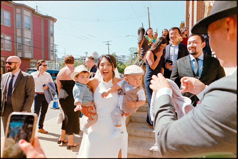 Joyful bride in white dress holds two babies while surrounded by cheering wedding guests outside.
