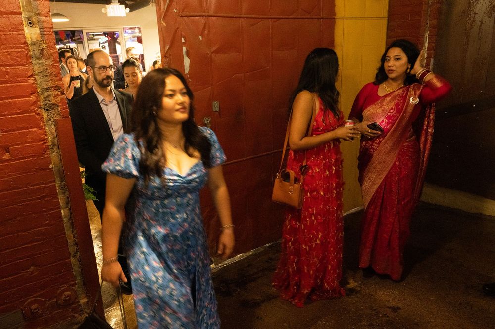 Group of people in elegant attire, including colorful saris, socializing in a warmly lit venue corridor.