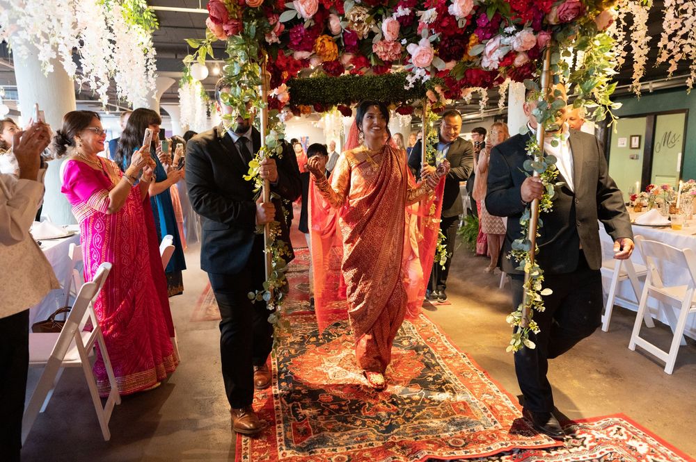 A bride in a red sari walks down the aisle under a floral canopy at a traditional Hindu wedding ceremony.