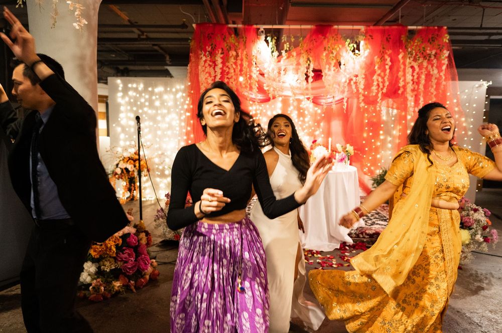 Women in colorful outfits laugh and dance joyfully at a vibrant Indian wedding reception celebration.