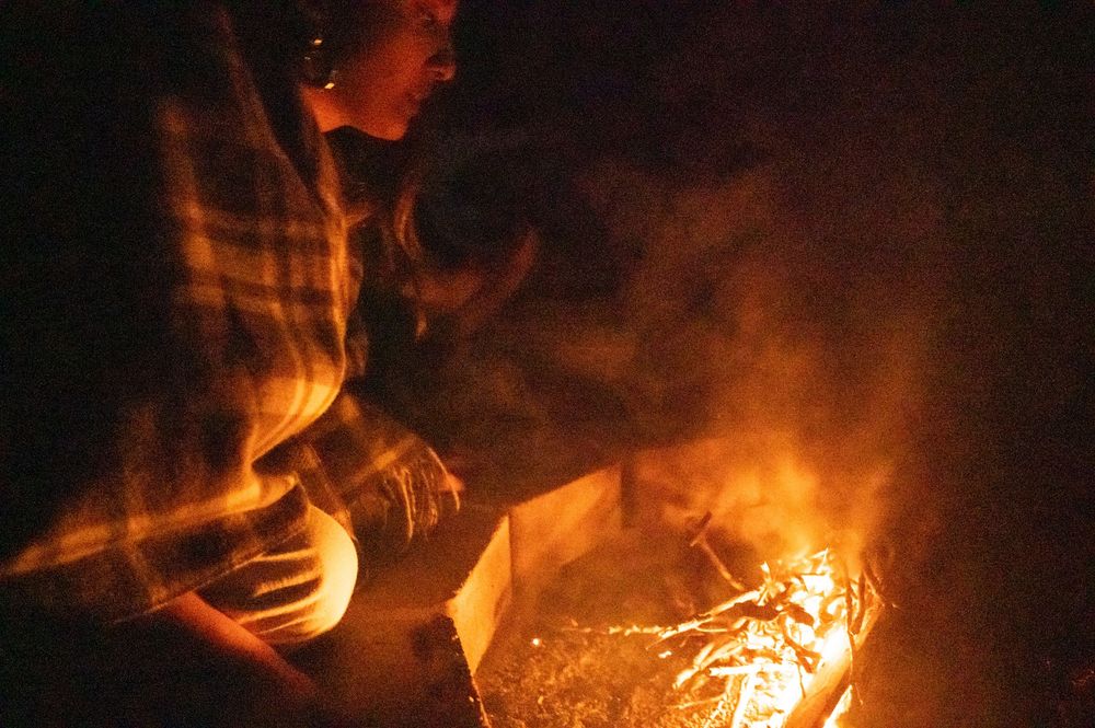 Person tending to a blazing campfire at night with smoke and flames glowing in the dark.