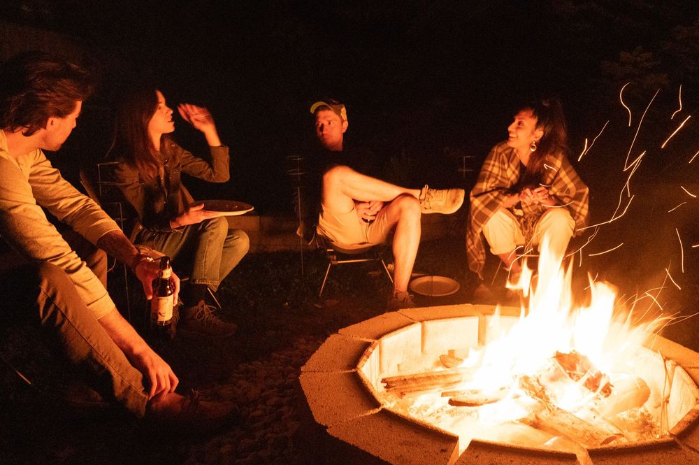 Group of friends sitting around a glowing campfire at night with sparks flying upward.