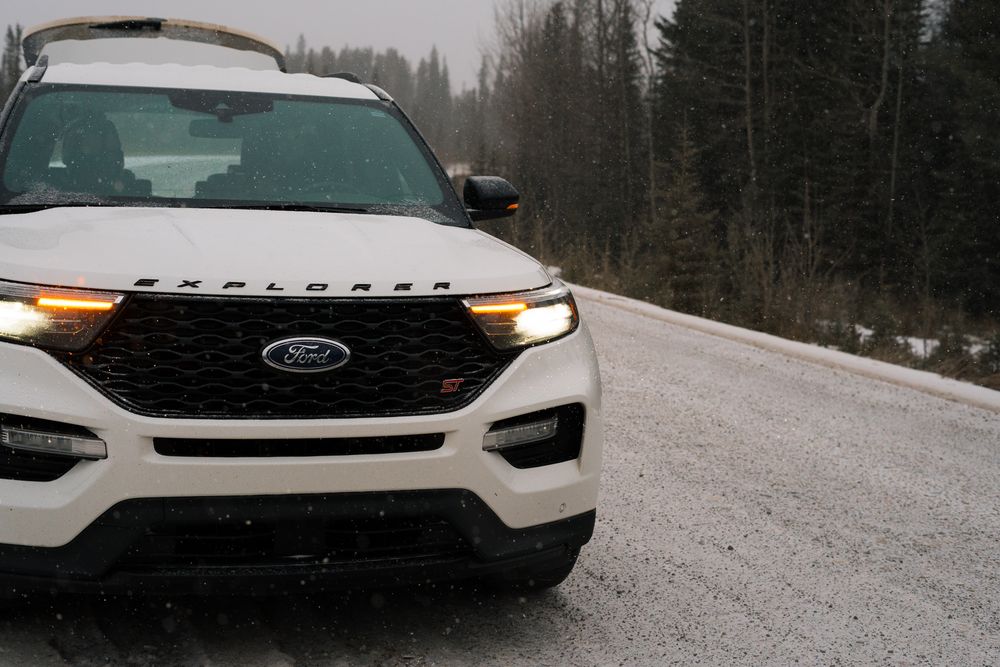 White Ford Explorer ST SUV with amber headlights on a snowy winter forest road in overcast conditions.