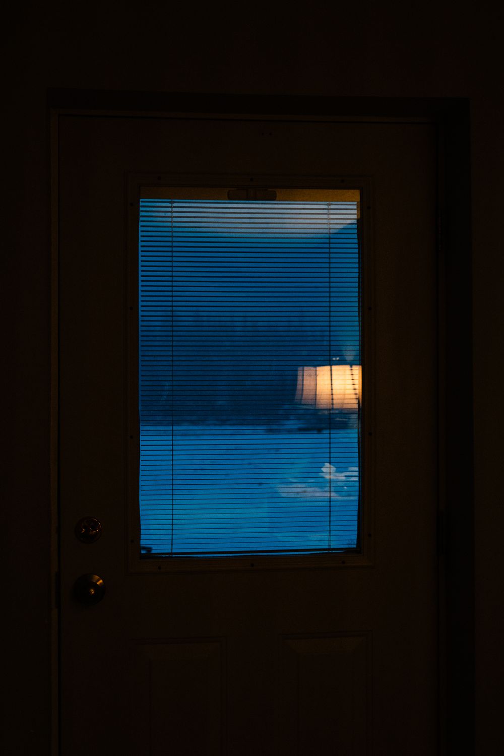 A dimly lit door with window blinds reveals a glowing lamp casting warm light in a dark blue room.
