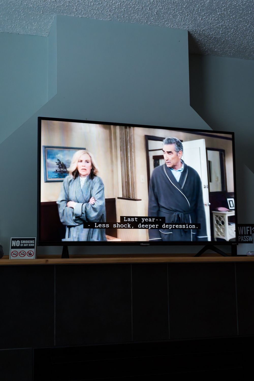 Flat screen TV displaying Schitt's Creek with subtitles, mounted on a dark wood entertainment stand in a dimly lit room.