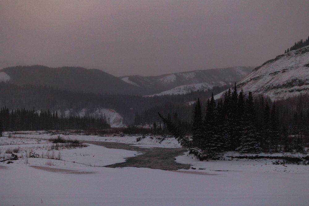 Snowy winter wilderness landscape at dusk with frozen river, evergreen trees, and mountain backdrop.
