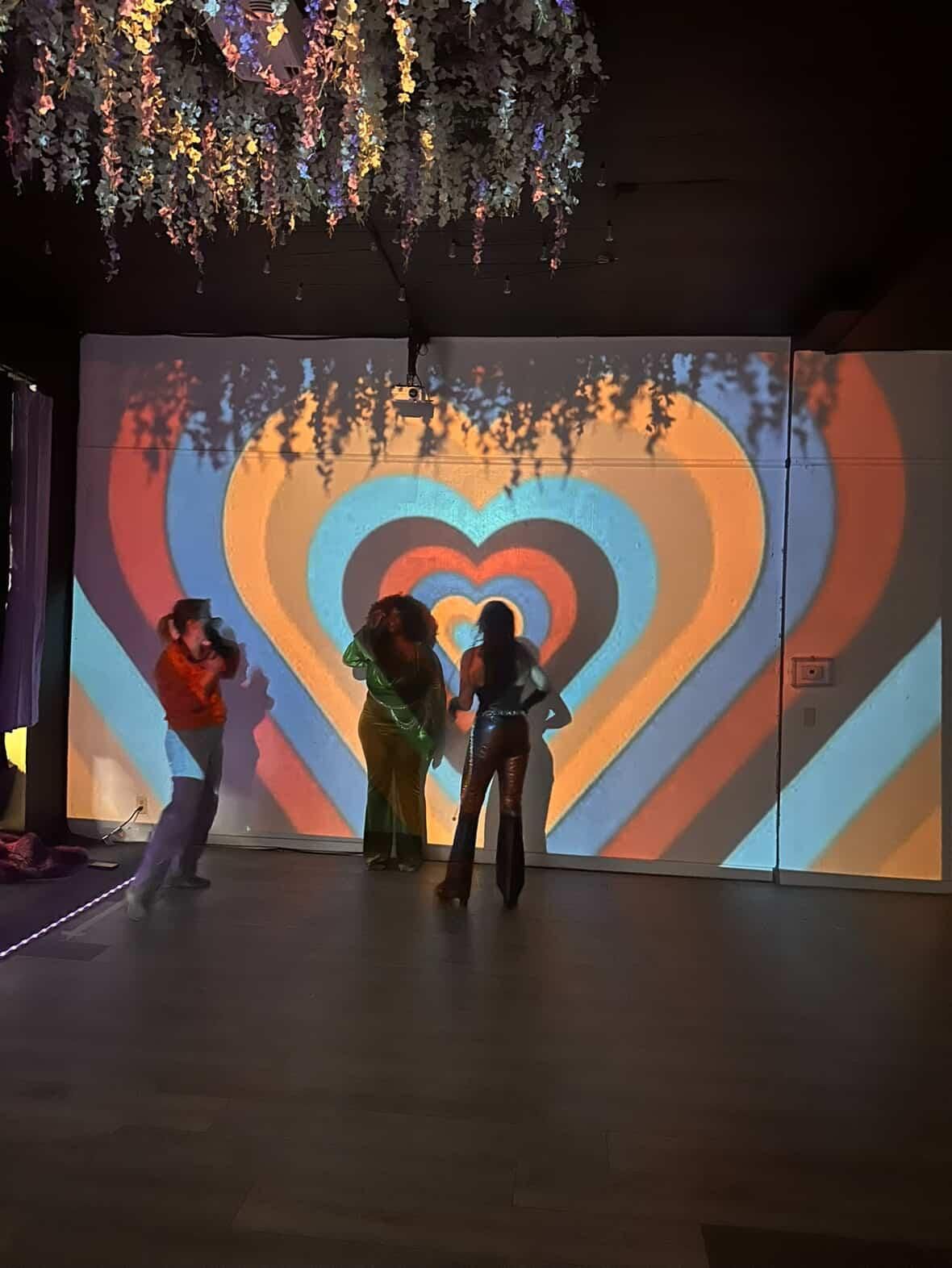 Silhouettes of people against a colorful geometric heart-shaped projection on a dark wall with hanging decorative lights.