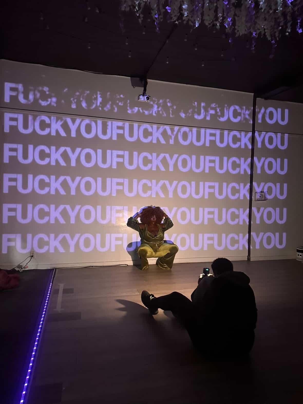 A sequence of projected text on a dark wall showing profanity repeated multiple times with silhouettes visible below.