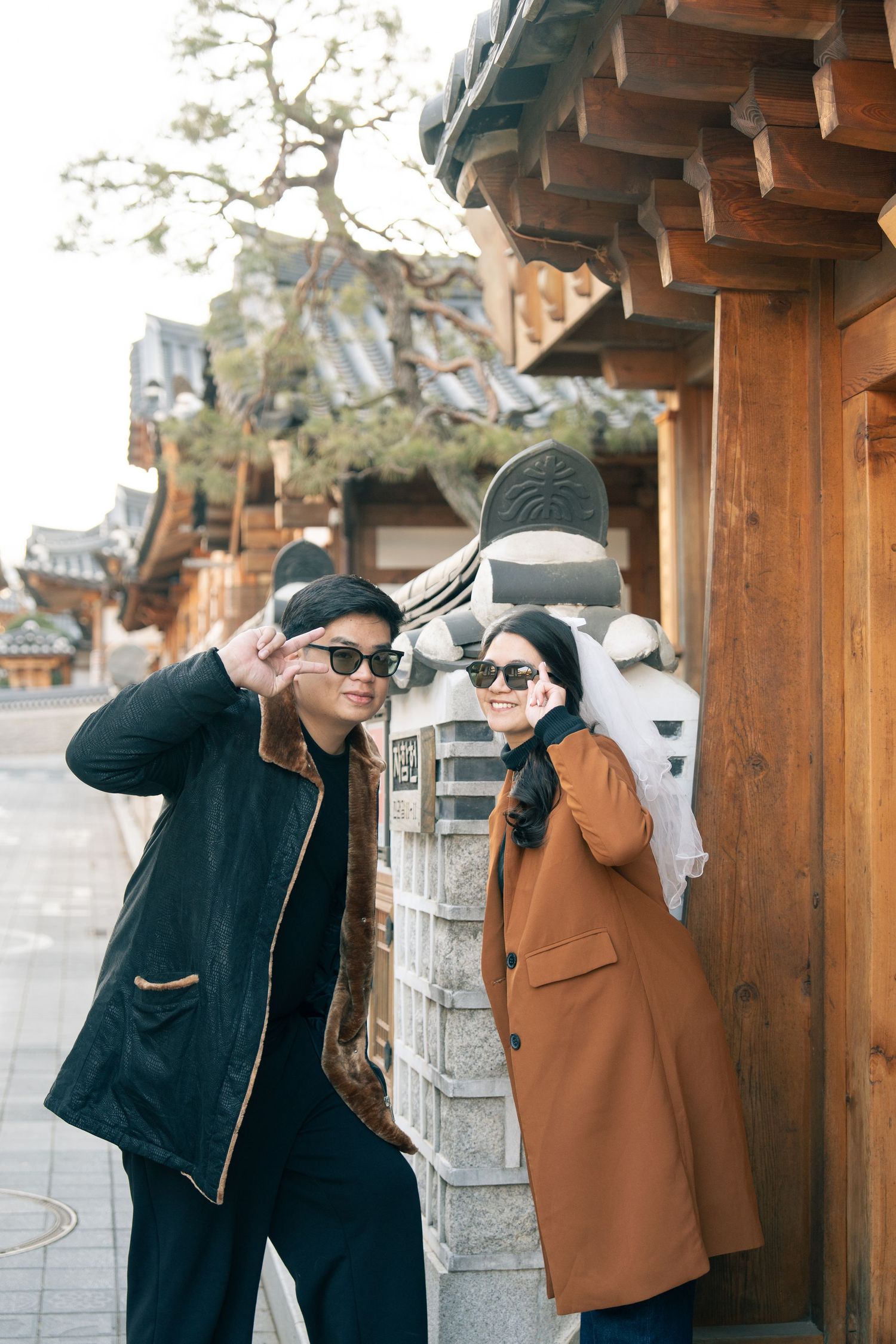 Two people in winter coats pose playfully in front of traditional Asian architecture on a bright day.