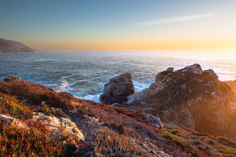 Dramatic coastal sunset over Big Sur with waves crashing against rocky cliffs and golden light illuminating the coastline.