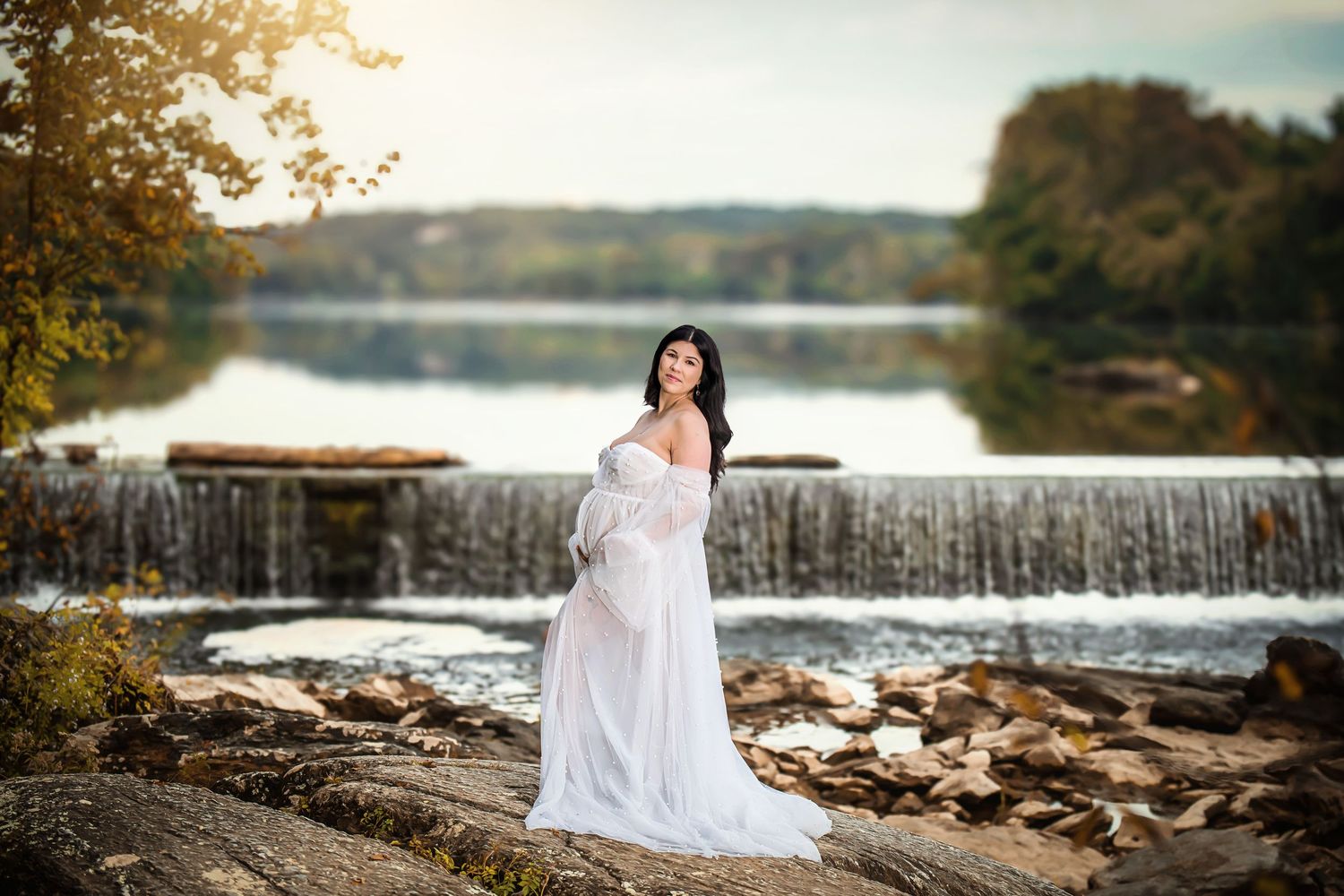 A flowing white dress contrasts against a scenic waterfall backdrop during golden hour at a serene lake setting.