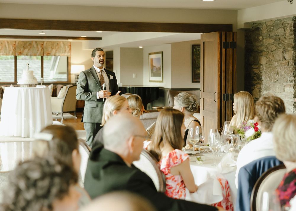 Wedding guest delivers heartfelt speech to seated guests during elegant indoor wedding reception dinner.