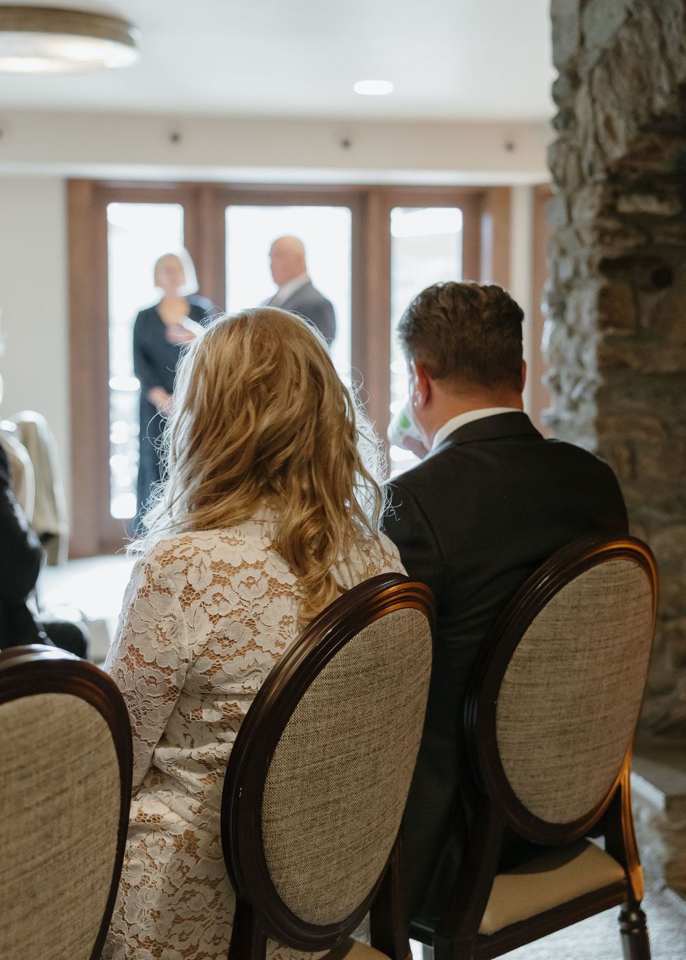 Bride in white lace dress and groom seated in ornate chairs during intimate wedding reception.