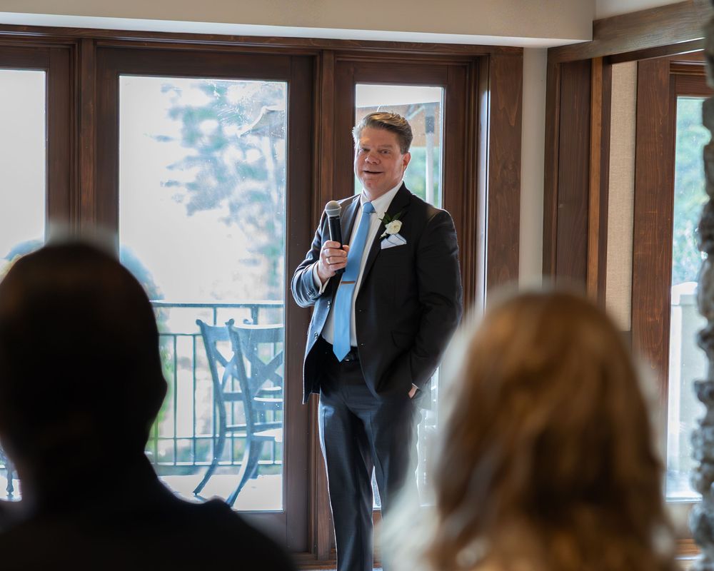 Groom in navy suit with boutonniere speaks confidently to wedding guests during reception celebration.