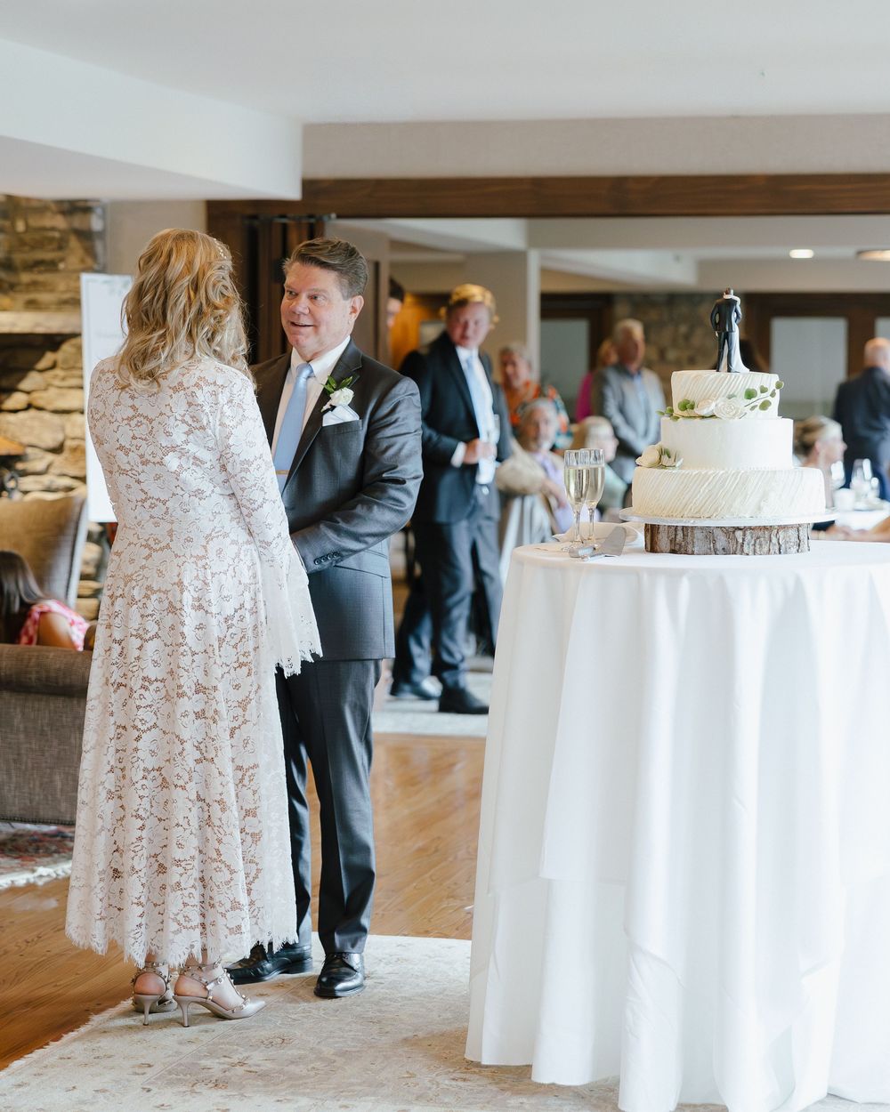 Bride in lace dress and groom in gray suit share a moment near wedding cake at reception.