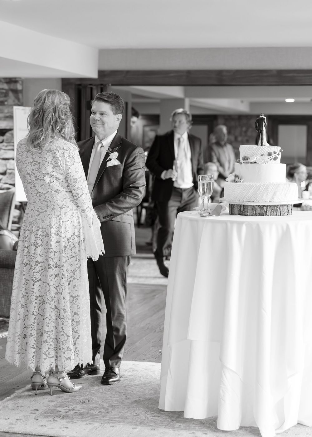 Black and white photo of bride and groom smiling near tiered wedding cake at reception.