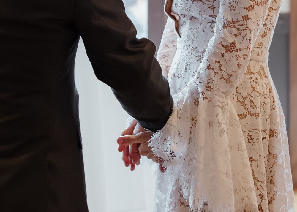 Close-up of bride and groom holding hands, bride wearing elegant long-sleeve lace wedding dress.