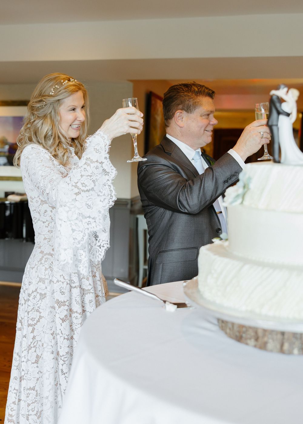 Smiling bride and groom toast with champagne glasses beside white tiered wedding cake.