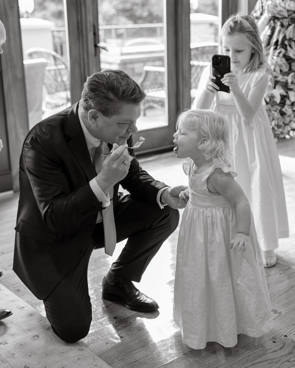 Black and white photo of groom kneeling to share cake with young flower girl at wedding.