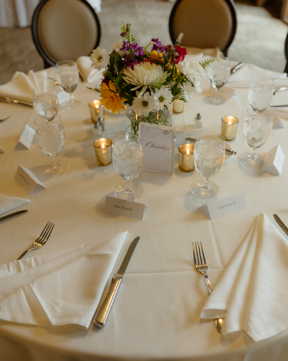 Elegant wedding reception table set with white linens, gold candles, crystal glasses, and colorful floral centerpiece.