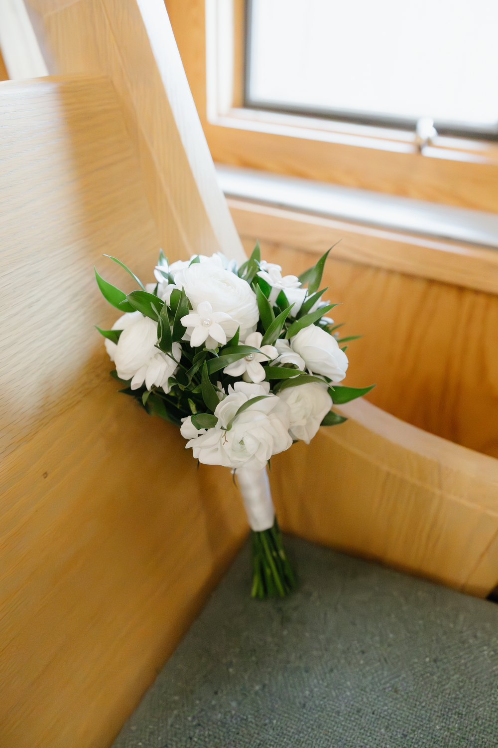 Elegant bridal bouquet of white roses and greenery resting on wooden church pew before wedding ceremony.